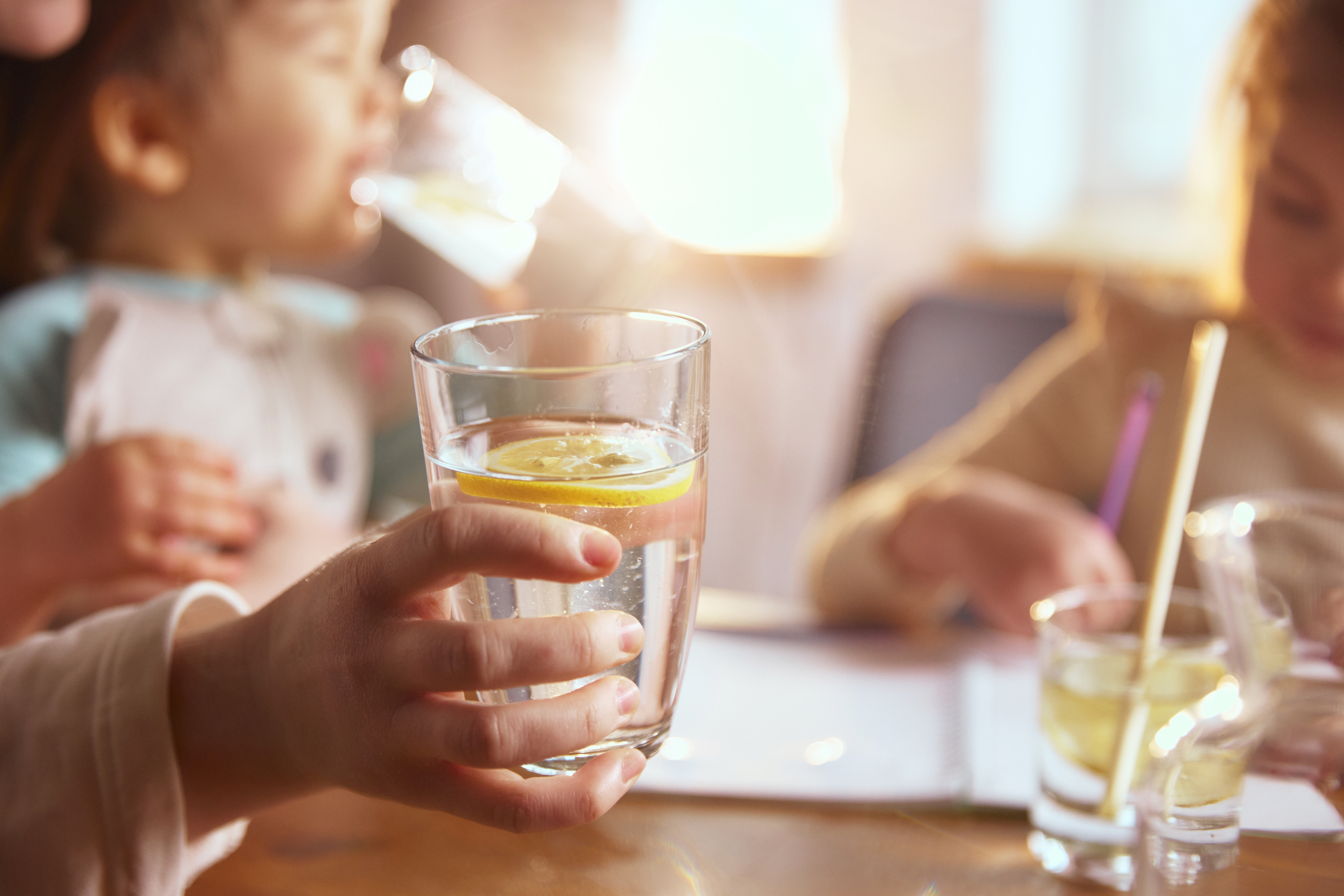 Ein Glas Wasser mit Zitrone, während die Kinder am Tisch zu Hause zeichnen. Das Konzept von sauberem Trinkwasser, gesunder Flüssigkeitszufuhr für Kinder, einfachen Alltagsgewohnheiten, familiärer Fürsorge und achtsamen Momenten im Alltag führen zu guter Trinkwasserhygiene.