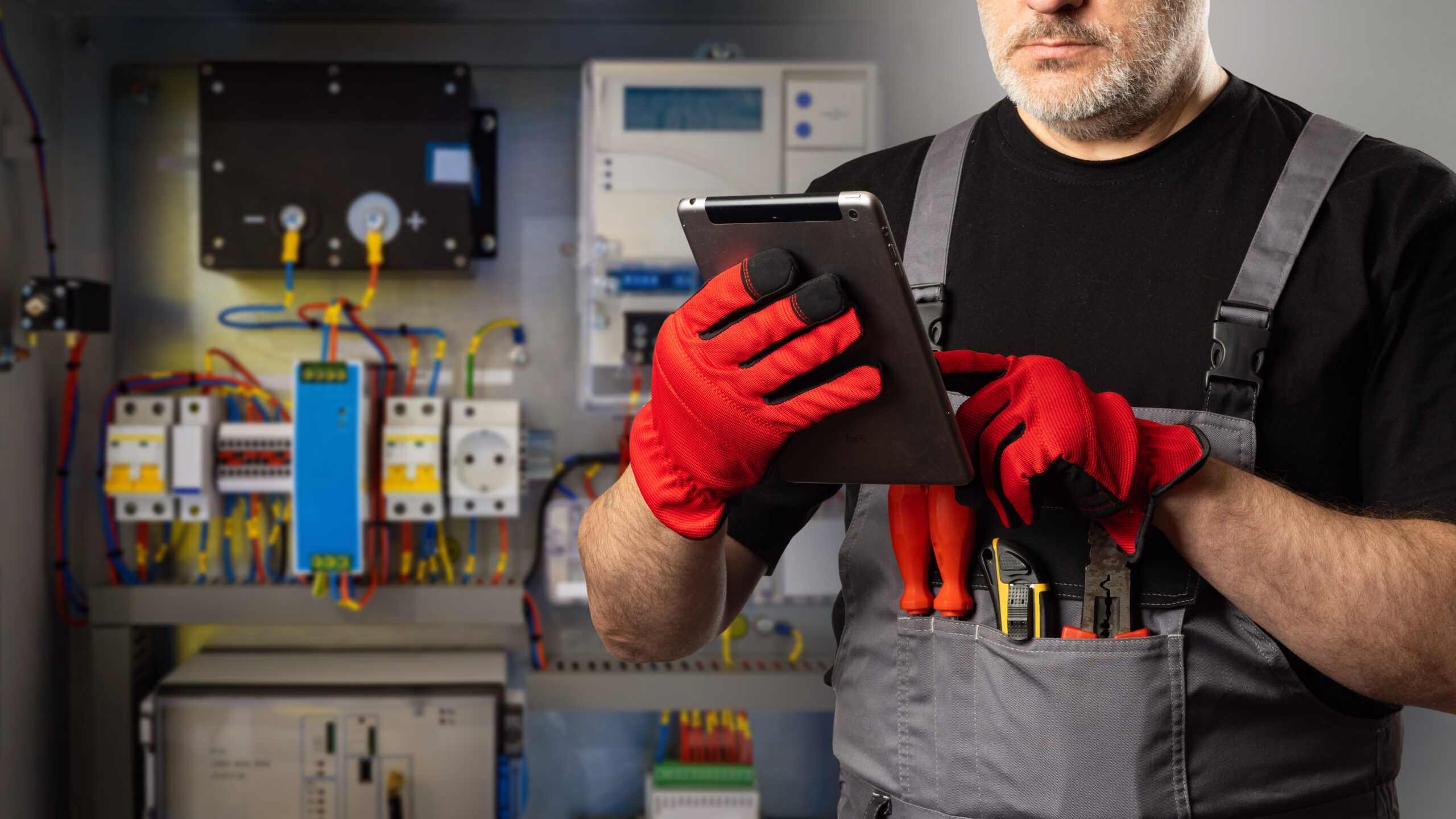 Man is electrician doing the E-Check. Electrical equipment installer. Electrician with tablet computer in his hands. Cropped wizard sets up automation system. Man electrician in gray overalls and gloves